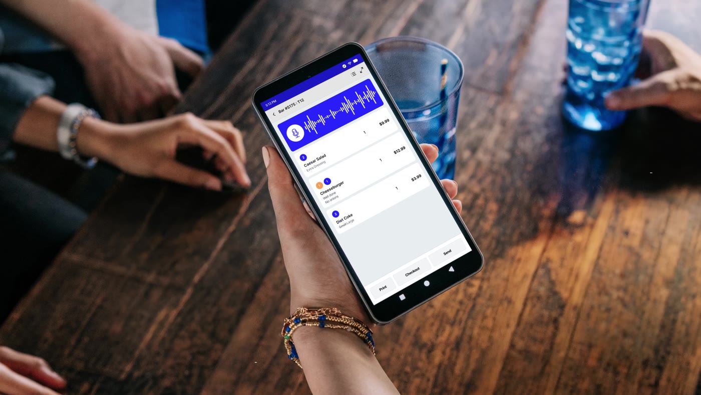 A server holding a Genius handheld at a table while an order builds itself on screen
